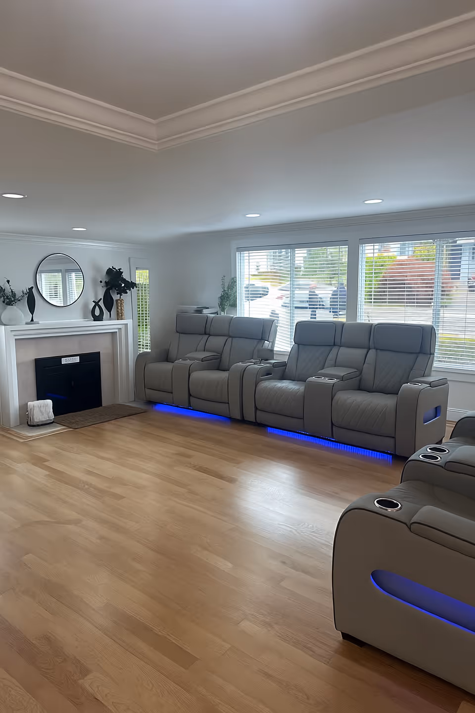 A modern living room with light wood flooring, three gray reclining chairs with blue LED lights underneath, a white fireplace with decorative vases and a round mirror above it, and large windows with white blinds letting in natural light.