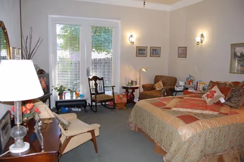A cozy bedroom with a large bed covered in a patchwork quilt, a wooden dresser with a lamp and framed photos, two chairs, a small table with plants near a glass door with blinds, and several framed pictures on the walls.