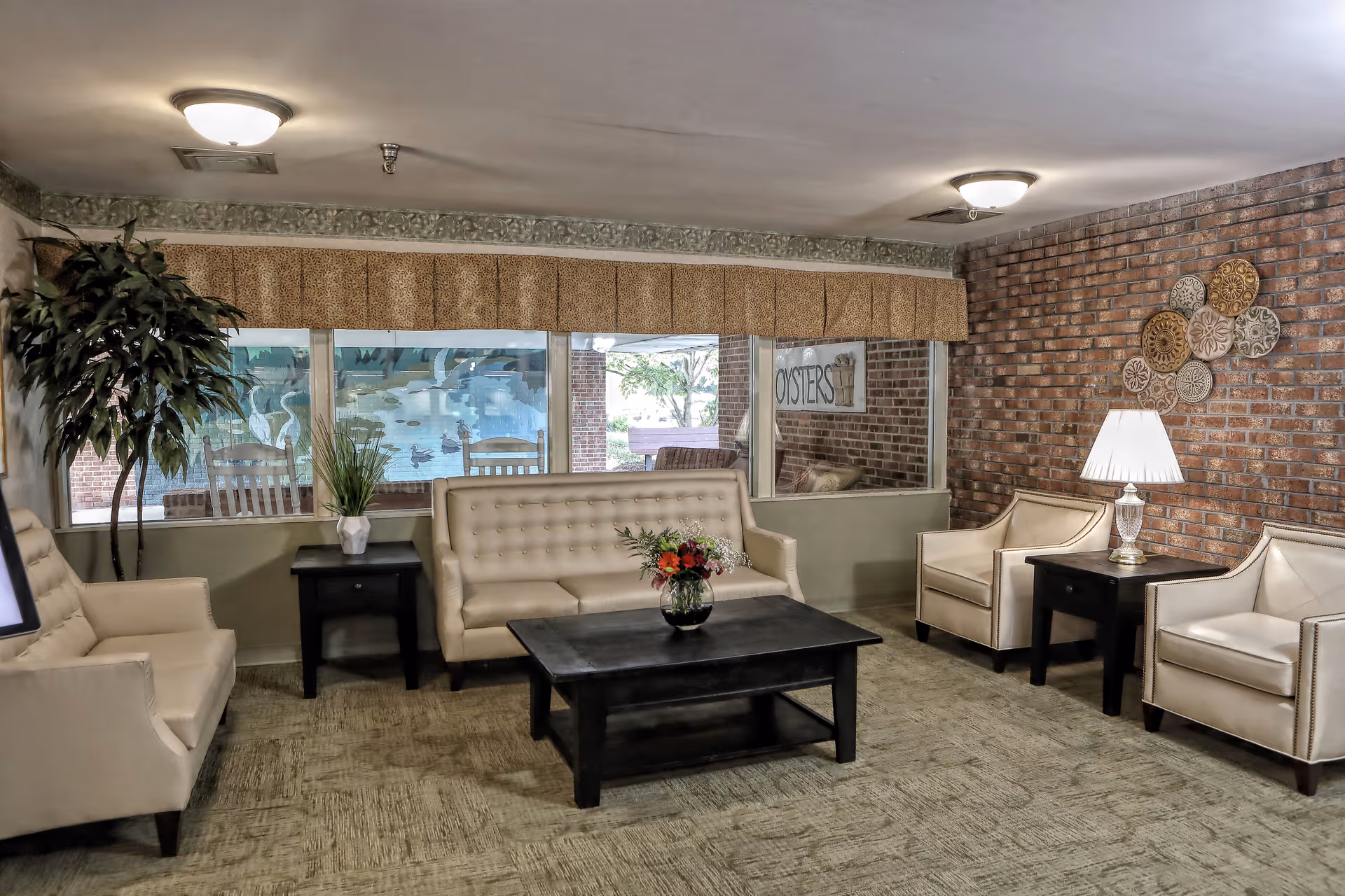 Communal living room with beige sofas and chairs around a dark coffee table, a brick accent wall, lamps, and large windows.