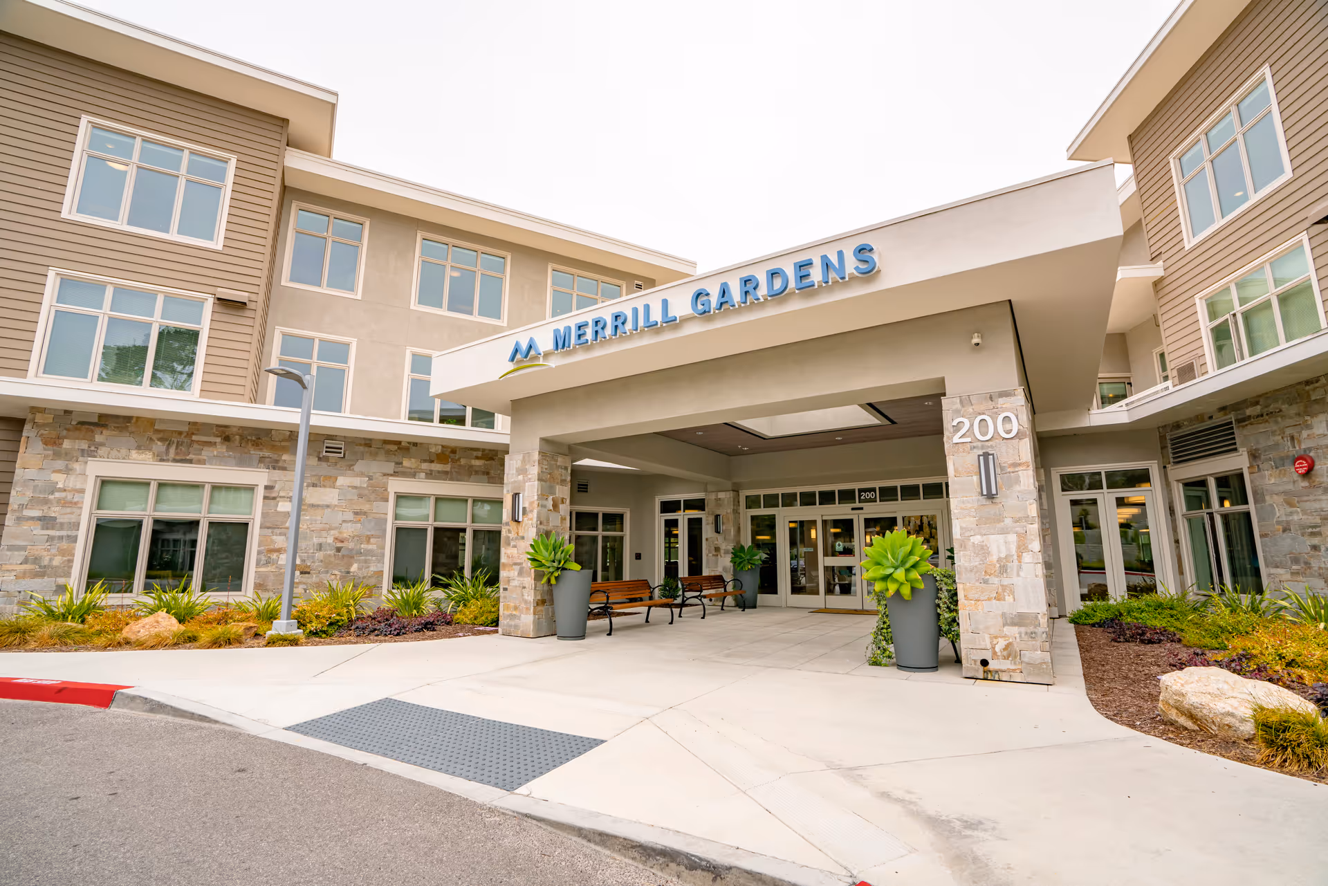 Front exterior view of Merrill Gardens at Monterey, showing the entrance with a covered drop-off area, benches, potted plants, and the building number 200 displayed on a stone pillar.