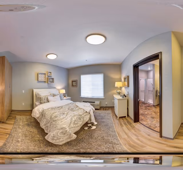 A cozy bedroom with a large bed covered in patterned bedding, two bedside tables with lamps, a dresser with a lamp and mirror, a window with blinds, wooden flooring, and an open doorway leading to a bathroom.
