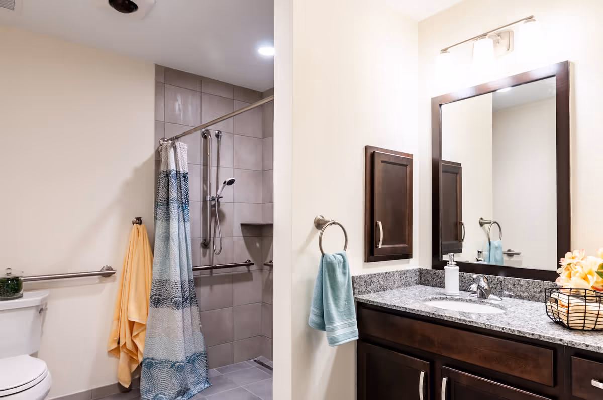 Well-lit modern bathroom with a granite vanity and mirror, sink, towels, toilet, and a tiled walk-in shower with a patterned curtain.