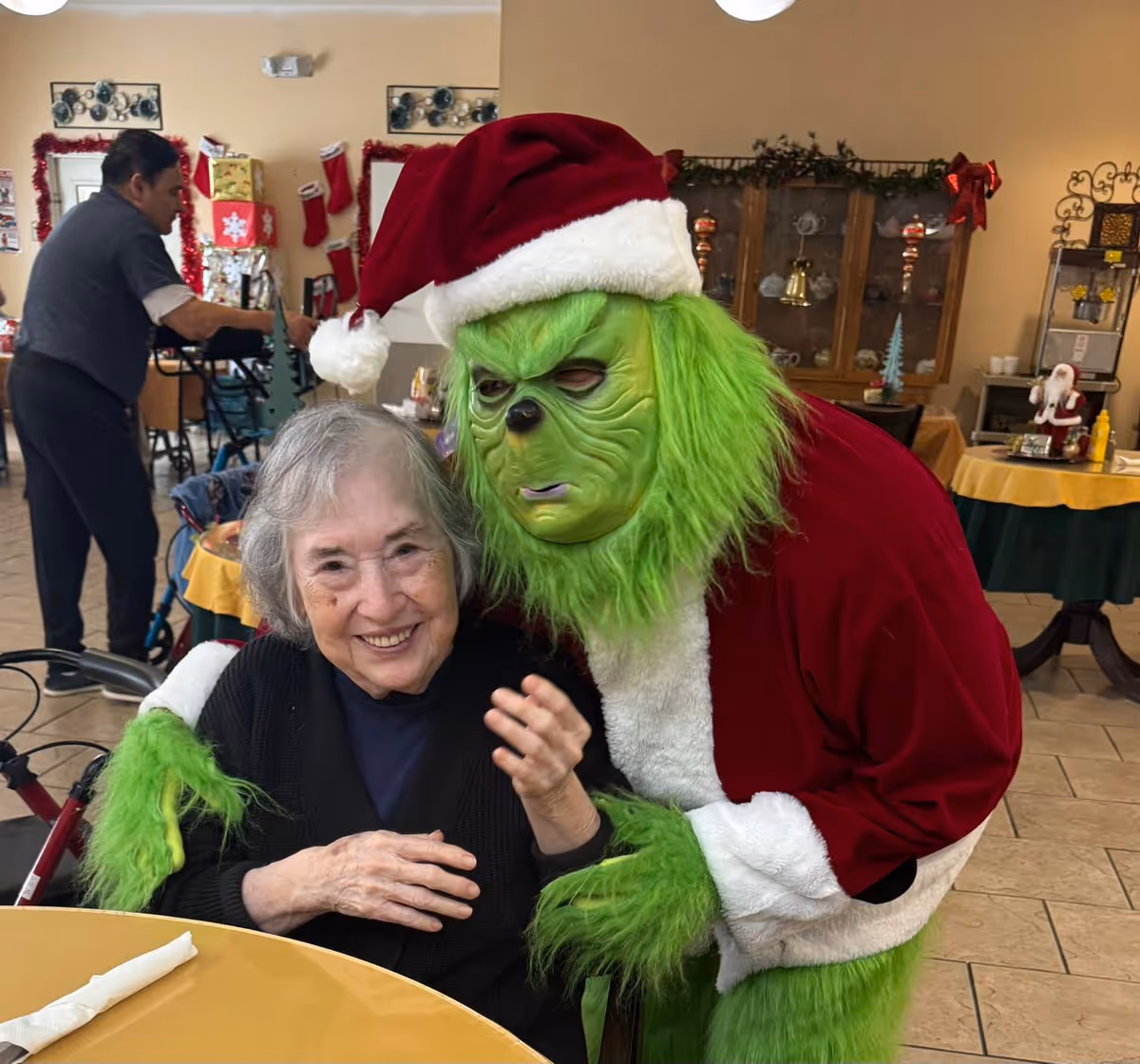 A person dressed as the Grinch in a Santa suit embraces a smiling elderly woman seated at a dining table in a decorated common room.