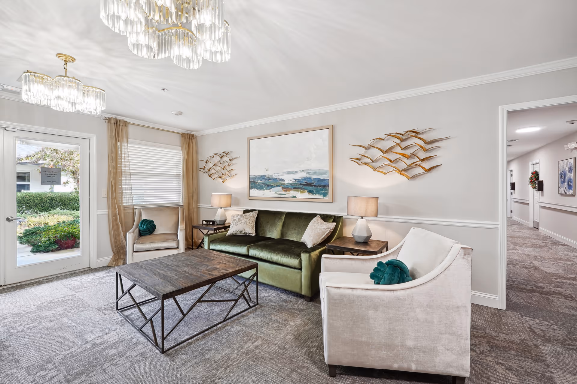 A bright and inviting living room area in a senior living facility with a green velvet sofa, two beige armchairs with green decorative pillows, a wooden coffee table with a metal frame, two side tables with lamps, and wall art including a large painting and metal bird sculptures. There is a glass door and window with sheer curtains letting in natural light, and a hallway visible to the right.