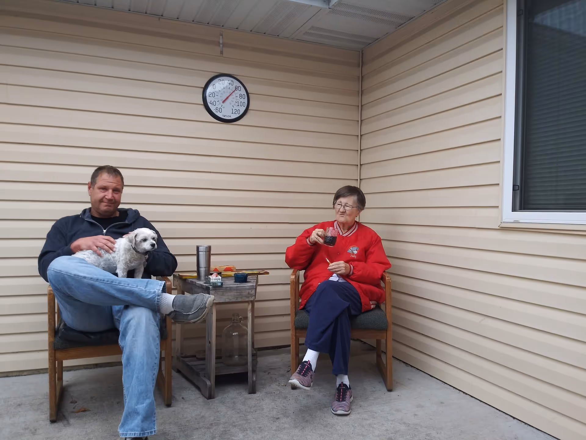 An elderly woman and a middle-aged man sitting on wooden chairs on a covered patio with beige siding walls. The man is holding a small white dog on his lap, and the woman is holding a drink. A small table with various items is between them, and a round thermometer is mounted on the wall above them.