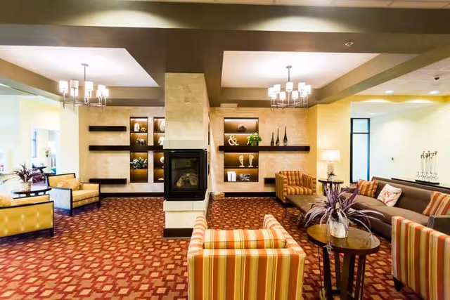 A spacious and well-lit living room area with patterned red carpet, multiple seating options including striped and solid armchairs, a long sofa, and a central fireplace with decorative shelves on either side. The room is decorated with plants, vases, and lamps, creating a cozy and inviting atmosphere.