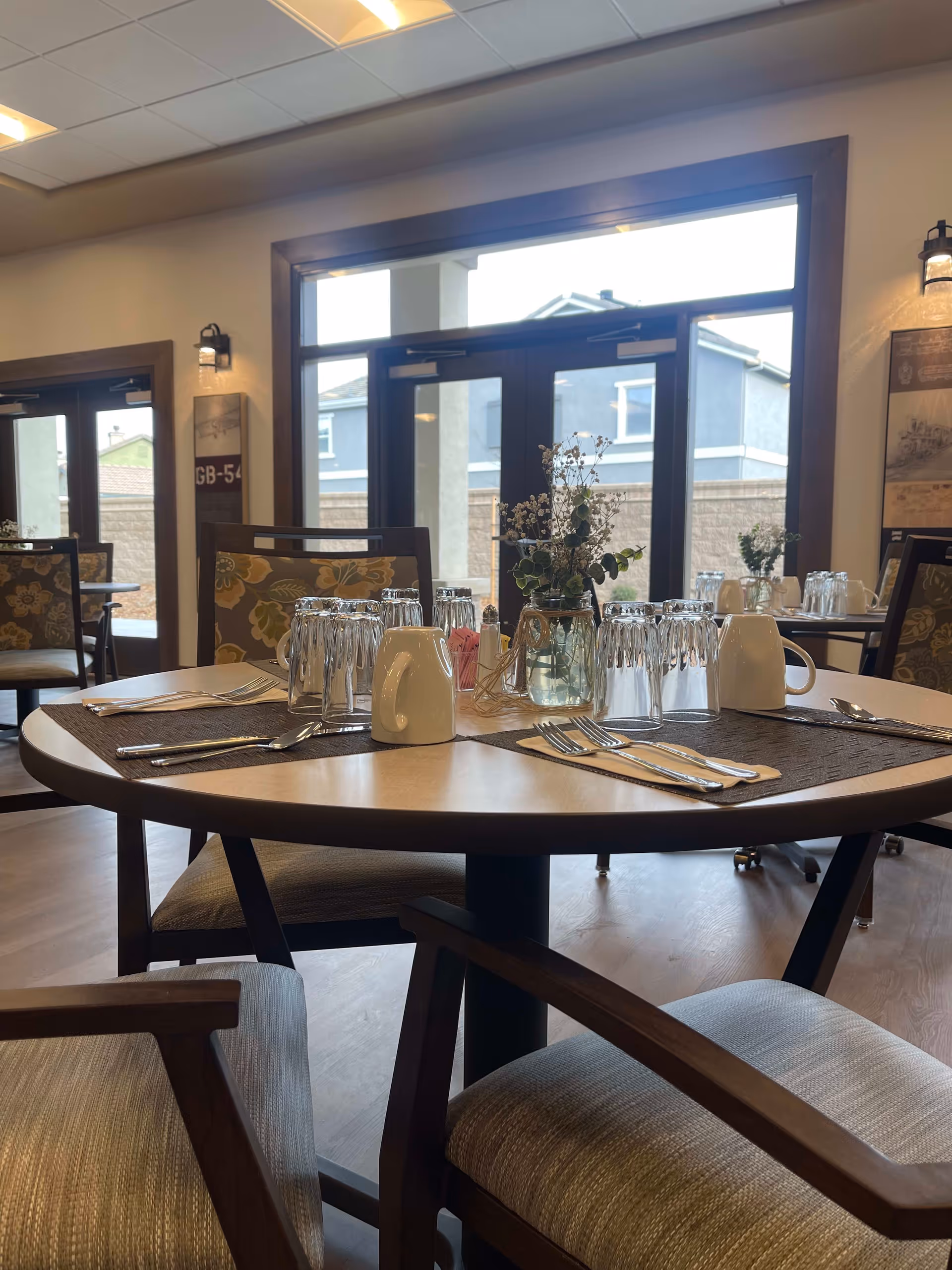 Interior view of a dining room with a round table set with upside-down glasses, white mugs, silverware on napkins, and a small vase with flowers in the center. The room has large windows and glass doors letting in natural light, with floral-patterned chairs around the tables.