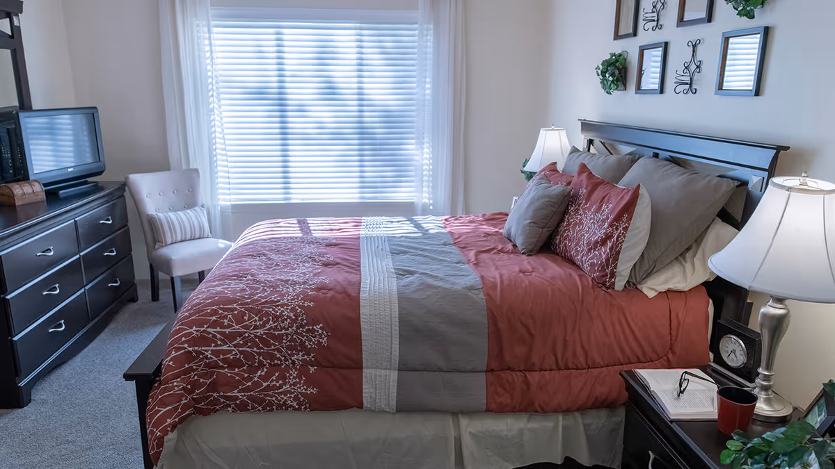 A well-lit bedroom with a large bed covered in a red and gray comforter with matching pillows. There is a nightstand with a lamp, clock, book, and a red cup. On the opposite side, there is a dresser with a TV on top and a white cushioned chair next to a window with closed blinds. The walls have decorative mirrors and small plants.