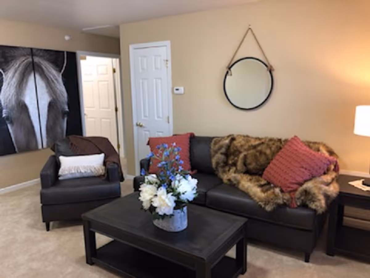 A cozy living room with a dark leather sofa adorned with a fur throw and two red pillows, a matching armchair with a white pillow and brown blanket, a black coffee table with a vase of white and blue flowers, a round mirror hanging on a beige wall, and a large black and white horse painting on the adjacent wall.