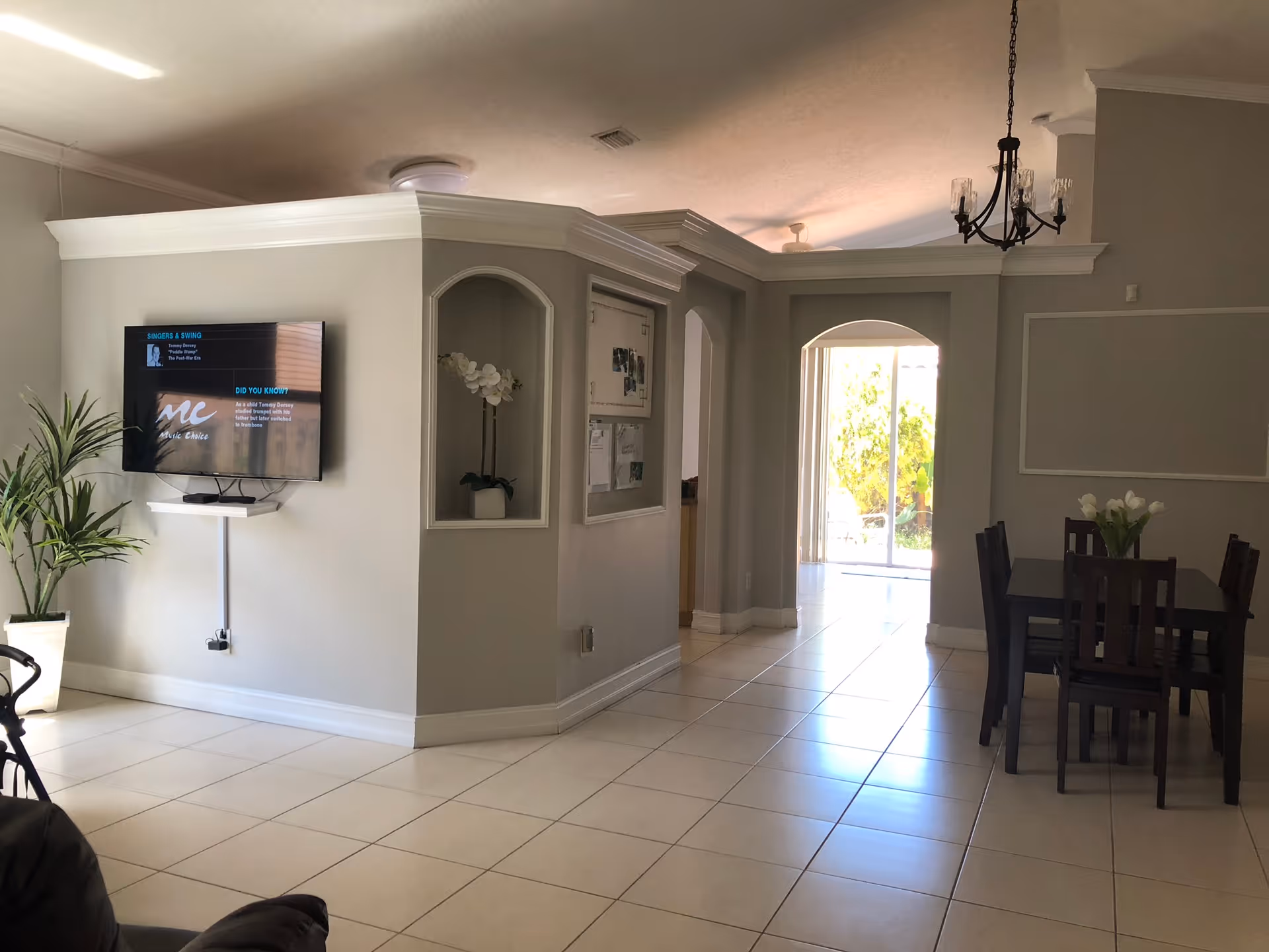 Bright open living and dining area with a wall-mounted TV, potted plants, and a dining table near an arched doorway leading outside.