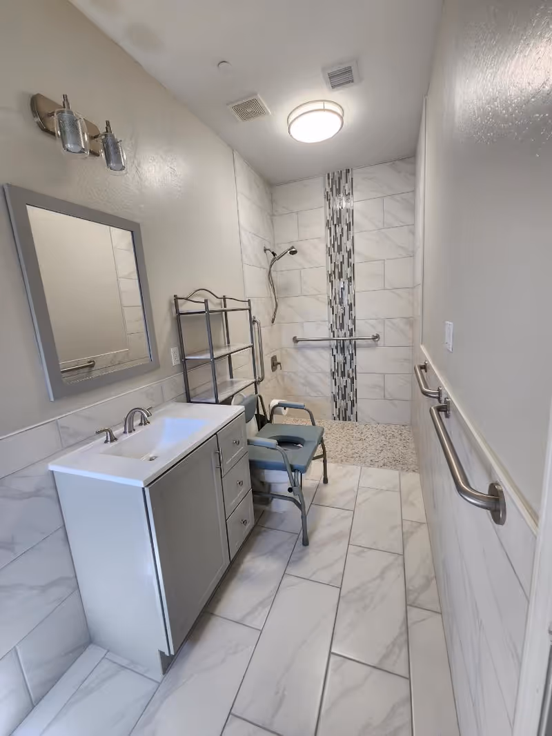A modern bathroom with white and gray marble tiles on the floor and walls. There is a gray vanity with a white sink and a mirror above it on the left side. A metal shelving unit and a shower chair are positioned near the walk-in shower area, which has a vertical strip of decorative tiles and a handheld showerhead. Stainless steel grab bars are installed on the walls for accessibility.