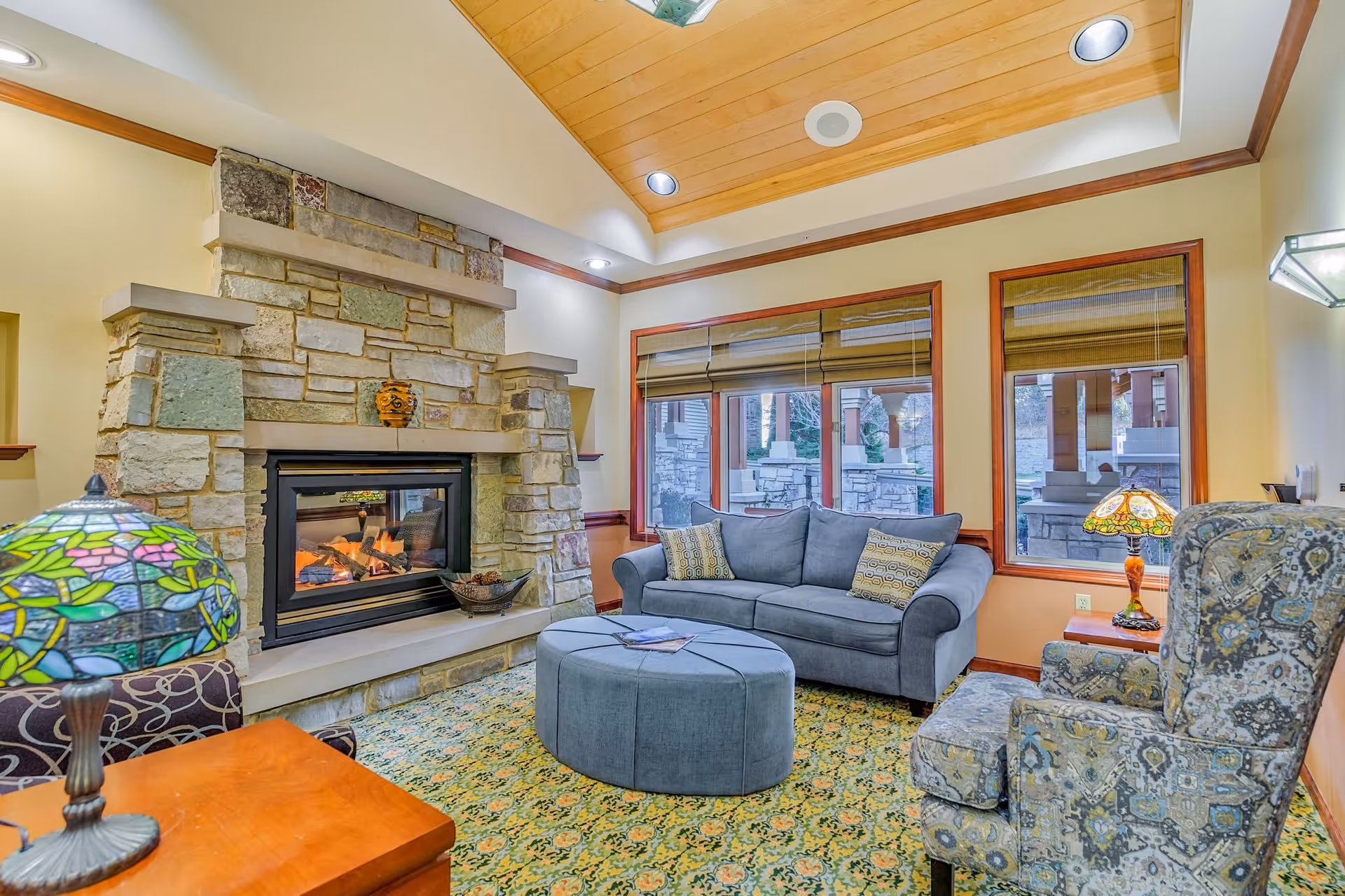 Cozy living room with a stone fireplace, a gray sofa with patterned cushions, a round ottoman, and a patterned armchair. The room has large windows with wooden frames and blinds, a colorful patterned carpet, and a wooden ceiling with recessed lighting. Two decorative table lamps with stained glass shades are visible.