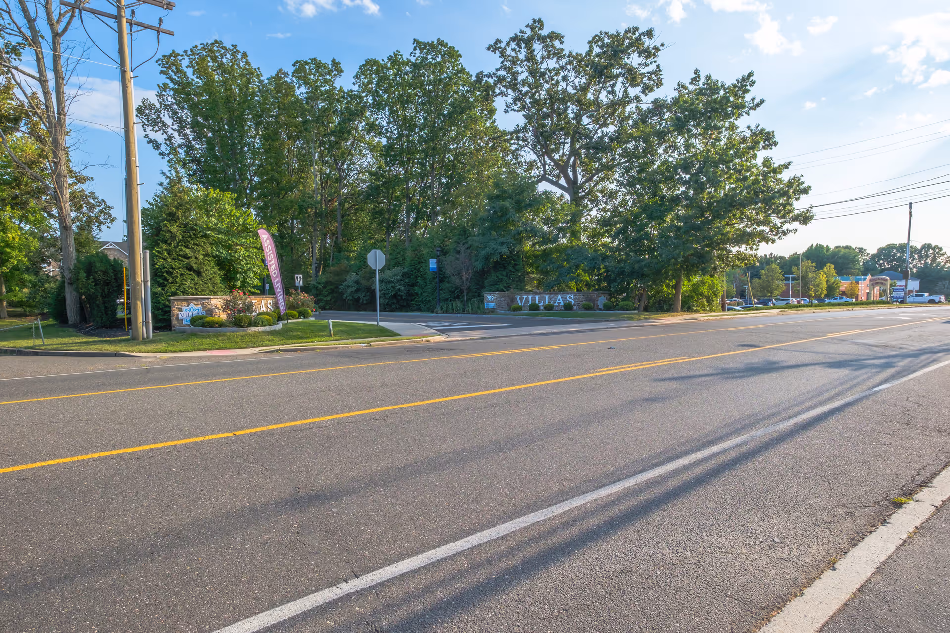 Wide view of a paved road and the tree-lined entrance sign for The Villas senior living community set back from the street.