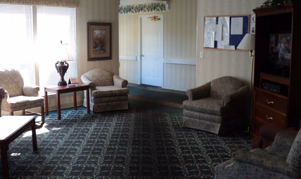 A cozy living room area with patterned carpet and several upholstered armchairs arranged around a wooden coffee table. There is a side table with a lamp and a framed picture on the wall. A television is visible on a wooden cabinet to the right, and a bulletin board is mounted on the wall near a white door.