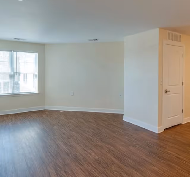 Empty room with wood flooring, beige walls, a white door on the right, and a large window on the left letting in natural light.