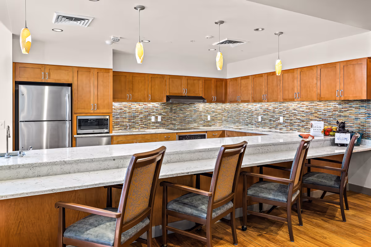A modern kitchen area with wooden cabinets, a stainless steel refrigerator, microwave, and dishwasher. The backsplash features a mosaic tile design in various earth tones. There is a long white countertop with four wooden chairs with cushioned seats arranged along it. Three pendant lights hang from the ceiling above the counter. On the counter, there is a small sign that says 'Welcome' and some decorative items.