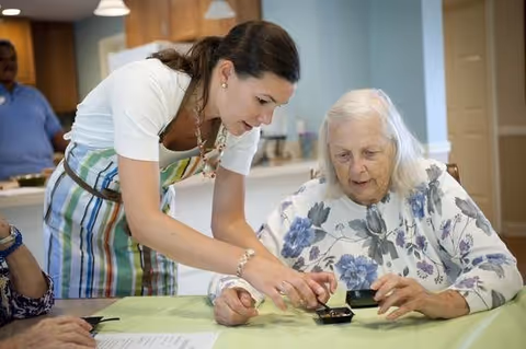 A young woman wearing a striped apron is assisting an elderly woman with white hair and a floral blouse at a table, showing her how to use a small device. Another person is partially visible sitting at the table, and a man stands in the background in a room with light blue walls and wooden furniture.