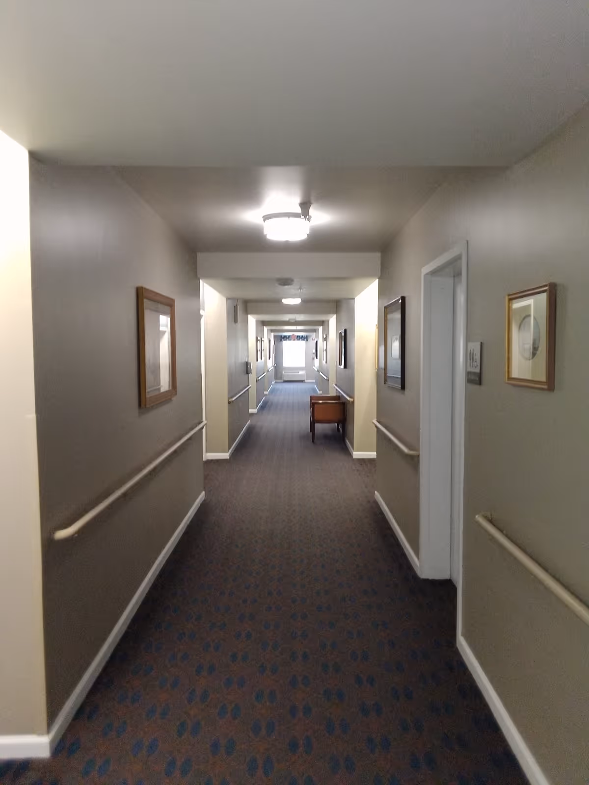 A long, carpeted hallway in a senior living facility with beige walls, handrails on both sides, framed pictures hanging on the walls, and ceiling lights. There is a small table along the right side of the hallway and a window at the far end letting in natural light.