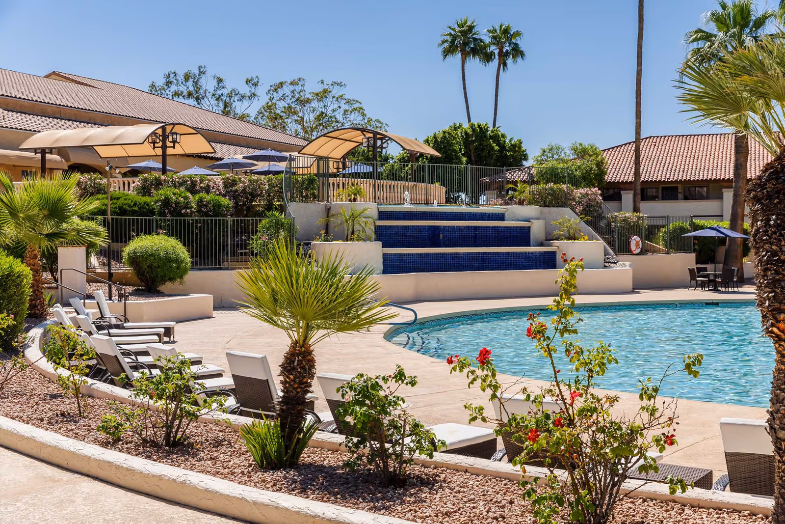 Outdoor swimming pool area with lounge chairs, palm trees, a tiered tiled water feature and shaded seating in front of red-tiled buildings.