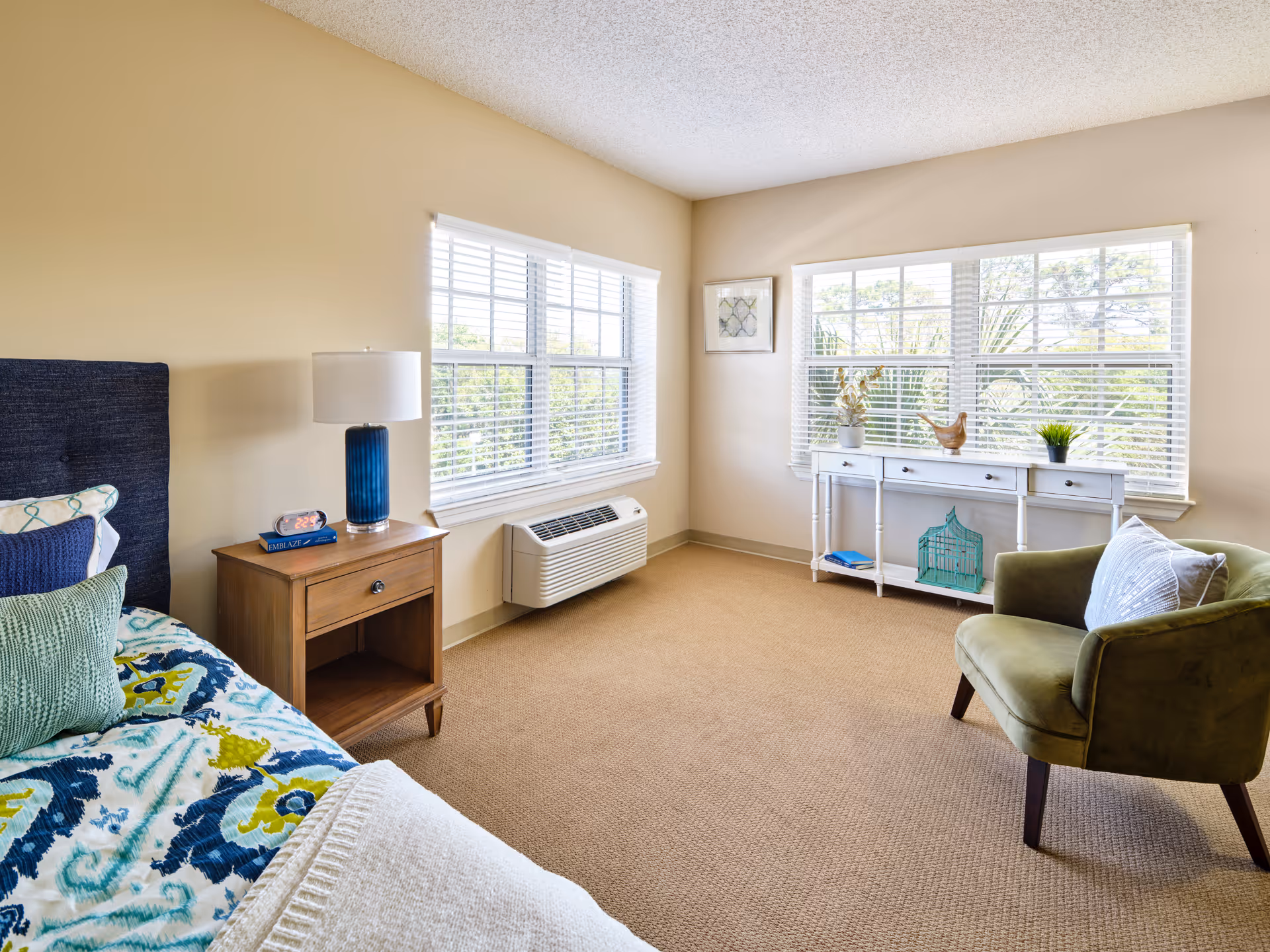 Bright furnished bedroom with a bed and nightstand, a green armchair, and large windows with blinds.