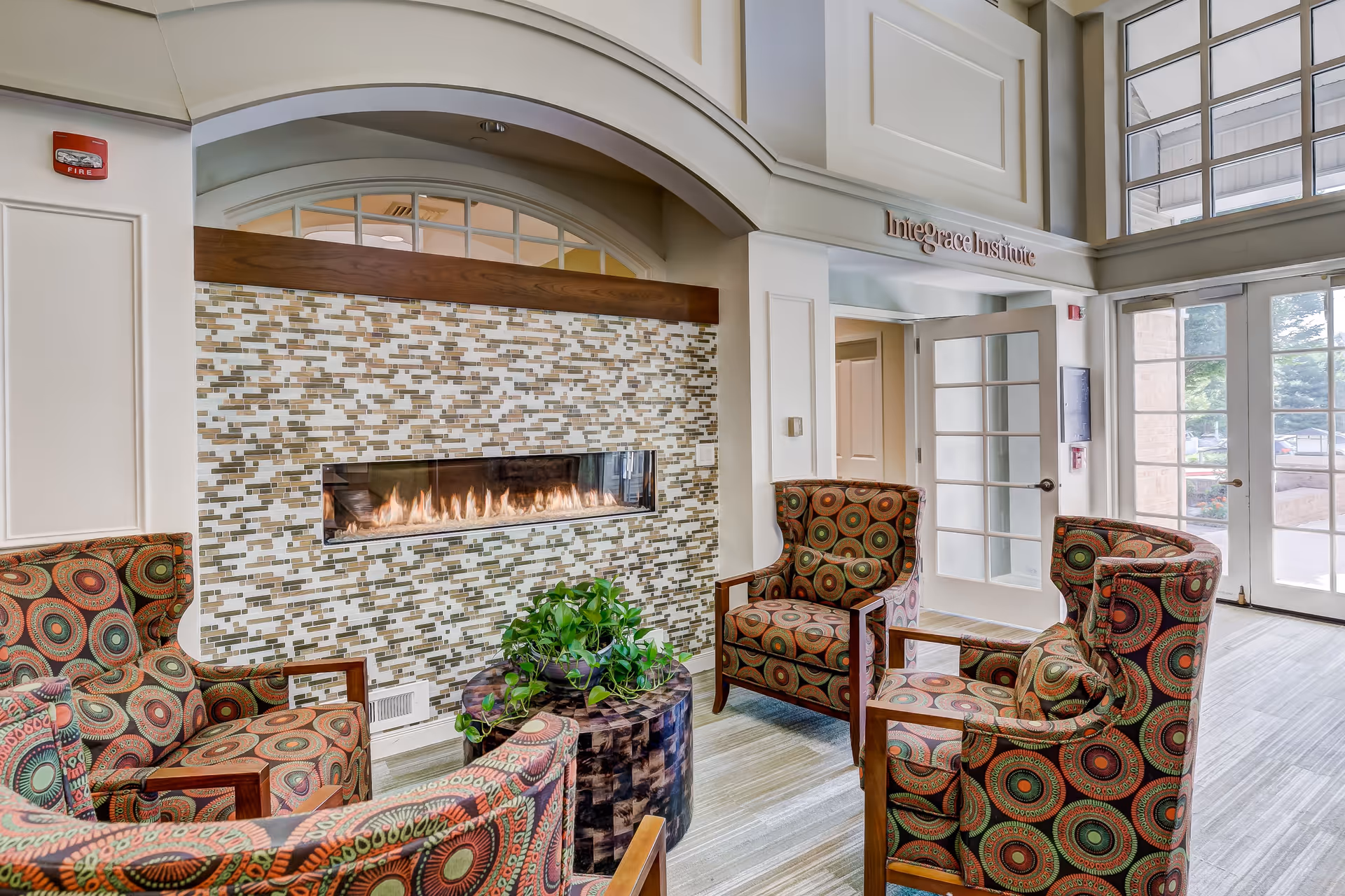 A cozy seating area with four patterned armchairs arranged around a small round wooden table with a green plant on it. Behind the chairs is a modern rectangular gas fireplace set into a wall with mosaic tile. The room has high ceilings, large windows, and a door leading outside. Above the door is a sign that reads 'InteGrace Institute.'