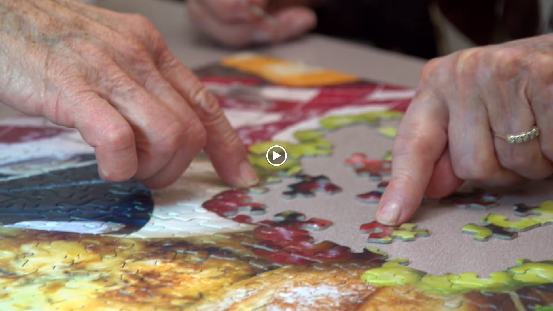 Close-up of two elderly hands working together to assemble a colorful jigsaw puzzle on a table.