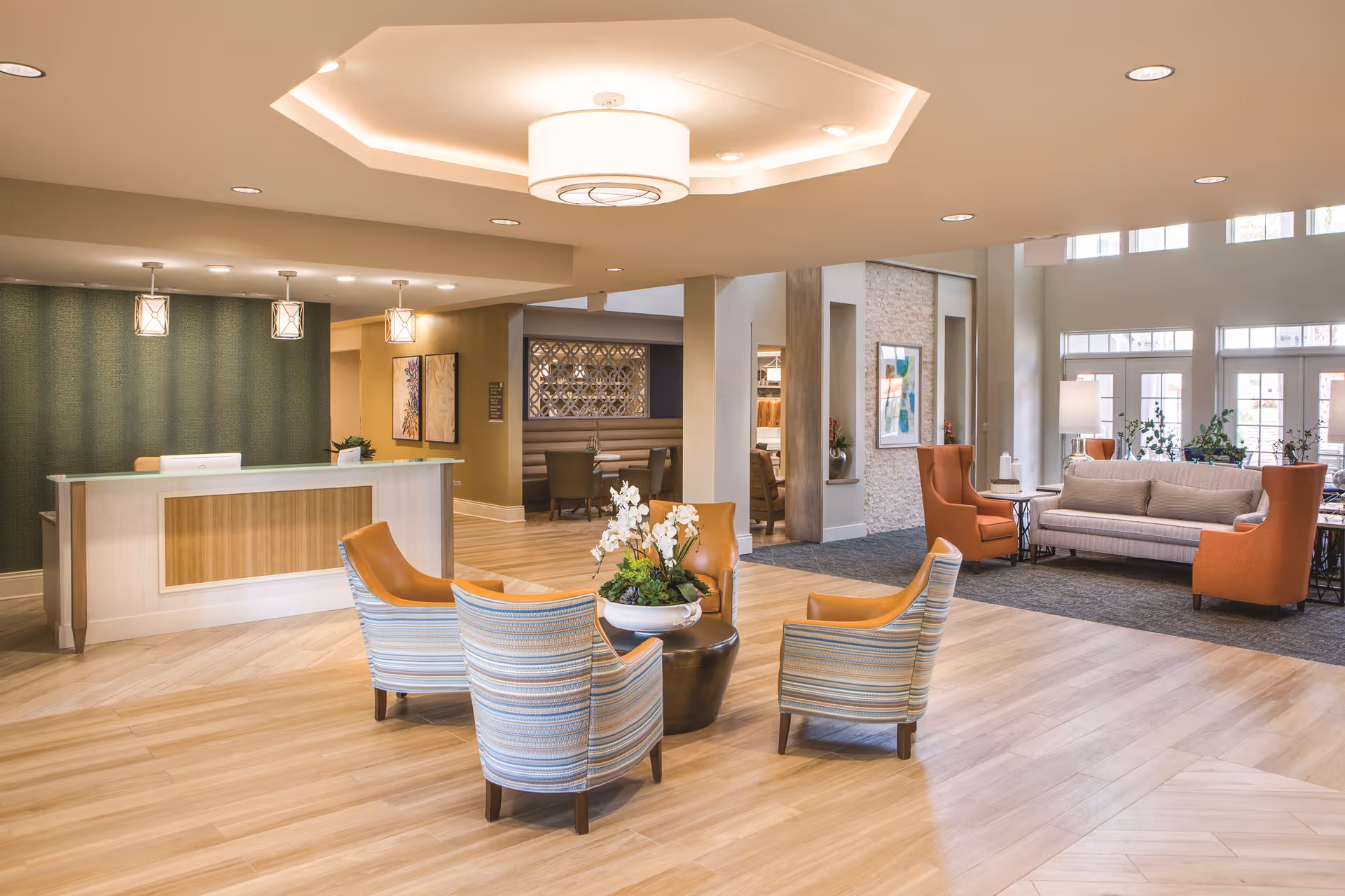 Spacious senior living lobby with a reception desk, grouped chairs around a central table, sofas, and large windows.
