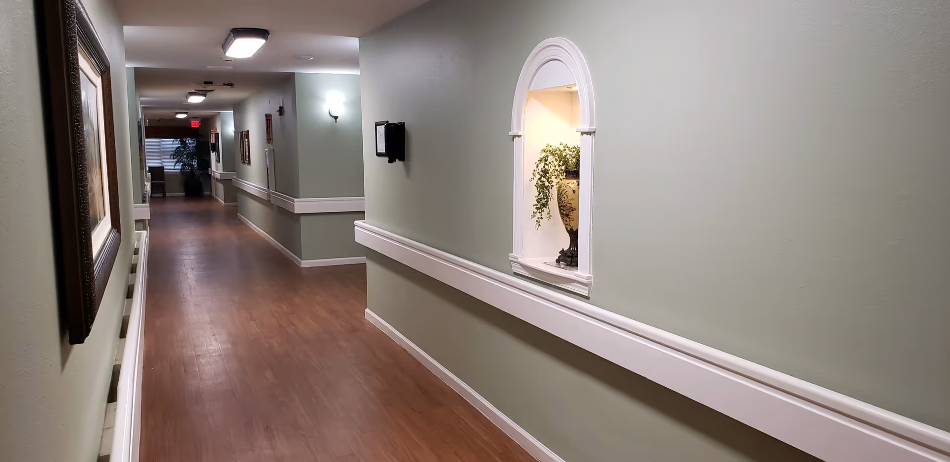 A long, well-lit hallway with light green walls and wooden flooring. The hallway features white handrails on both sides, framed artwork on the left wall, and a decorative niche with a vase and plant on the right wall. There are ceiling lights and a window with blinds at the far end.