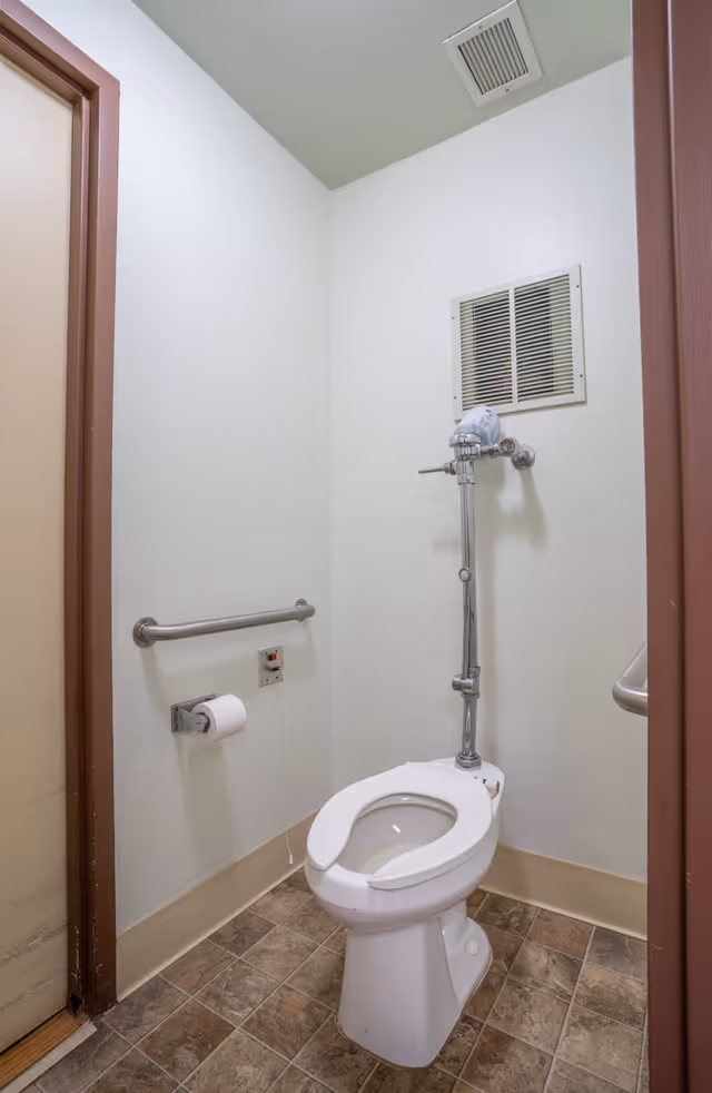 A small bathroom with a white toilet featuring a metal flush pipe. There is a metal grab bar on the wall to the left of the toilet, a toilet paper holder with a roll of toilet paper, and a ventilation grate above the toilet. The floor has brown and beige tiles, and the walls are painted white with a beige baseboard.