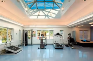 Bright fitness room with exercise equipment including a treadmill, stationary bike, and weight machine under a large skylight ceiling with windows showing outdoor greenery.