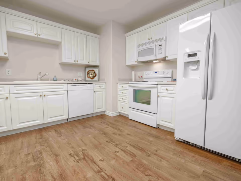 A clean kitchen with white cabinets, a white dishwasher, a white stove with an oven, a white microwave above the stove, and a white refrigerator with a water and ice dispenser. The floor is a wood-style laminate.