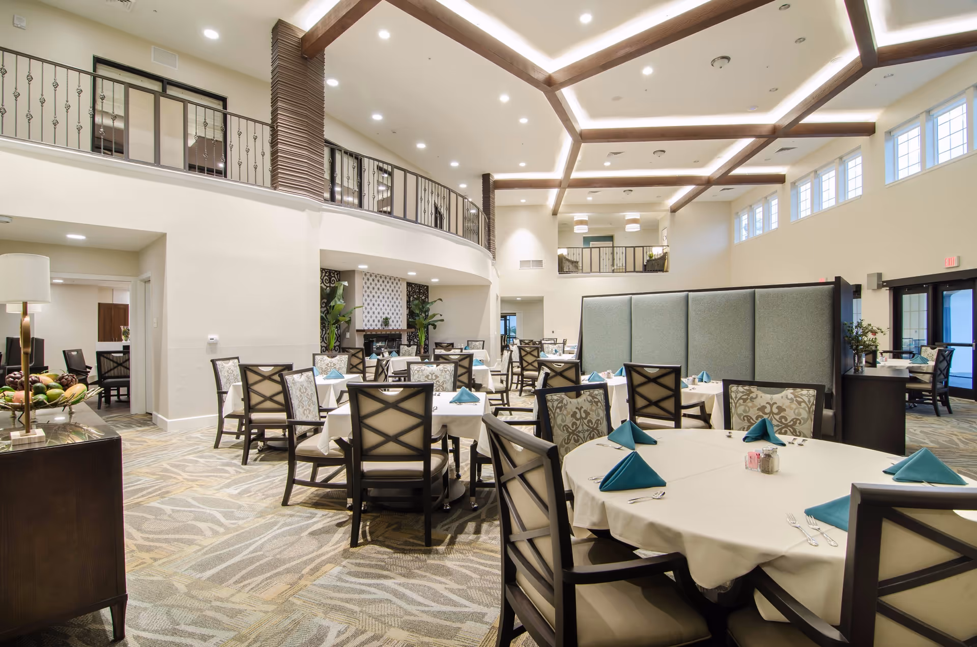 A spacious and well-lit dining room with multiple tables covered in white tablecloths, each set with teal folded napkins and silverware. The room features high ceilings with wooden beams and recessed lighting, large windows near the ceiling allowing natural light, and a second-floor balcony overlooking the area. There is a decorative fireplace with plants on either side and a patterned carpet covering the floor.