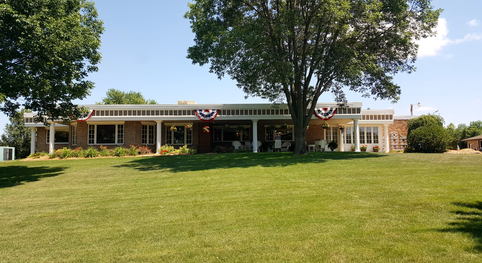 Single-story brick nursing home front with a wide grassy lawn, large trees, and a covered porch decorated with patriotic bunting.