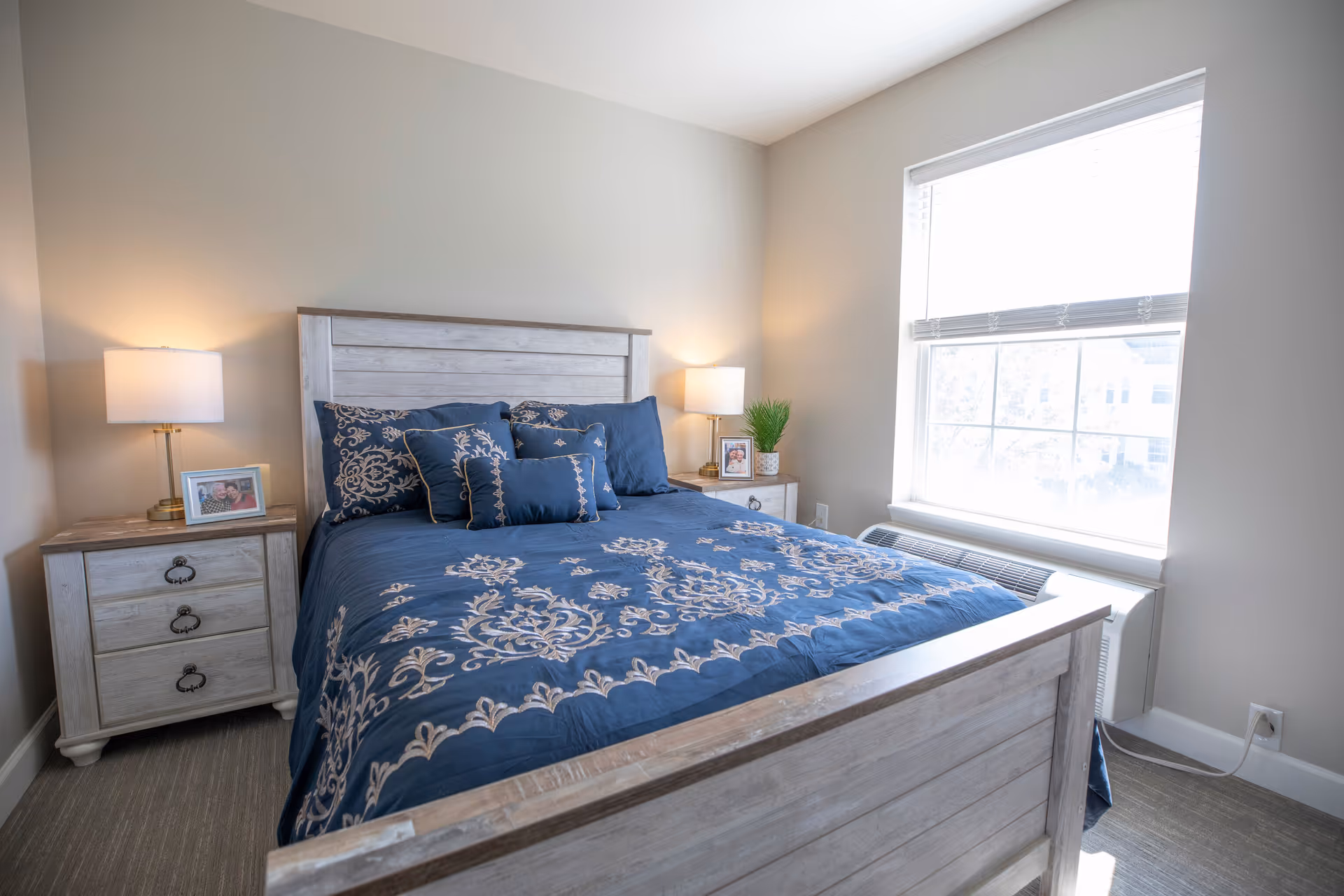 A bedroom with a wooden bed frame and matching nightstands on either side. The bed is made with a blue comforter and decorative pillows featuring a beige floral pattern. Each nightstand has a lamp and a framed photo. There is a large window with blinds letting in natural light, and a white air conditioning unit below the window.
