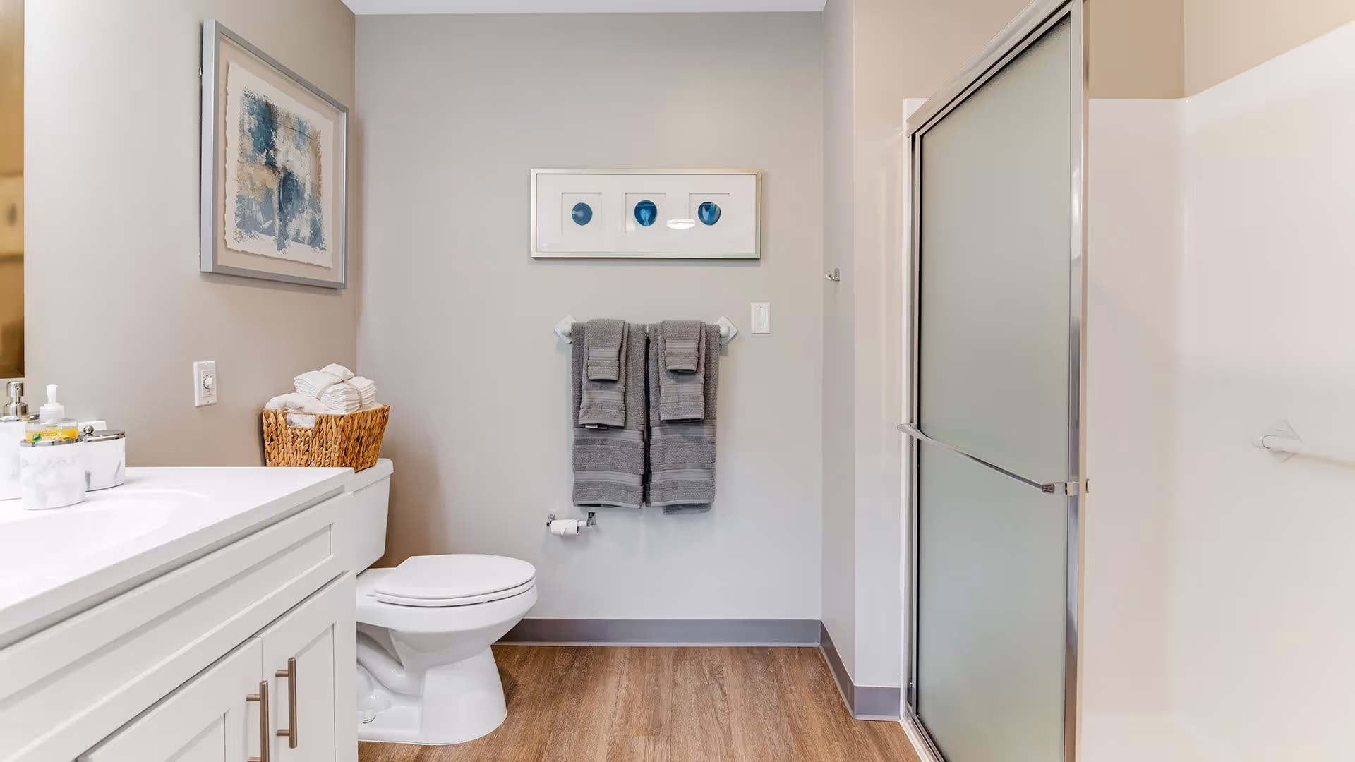 A clean and modern bathroom featuring a white toilet, a white vanity with a sink, a basket with rolled towels, two gray towels hanging on a wall-mounted towel rack, and a shower with frosted glass sliding doors. The walls are painted light gray and decorated with two framed abstract artworks. The floor has wood-style flooring.