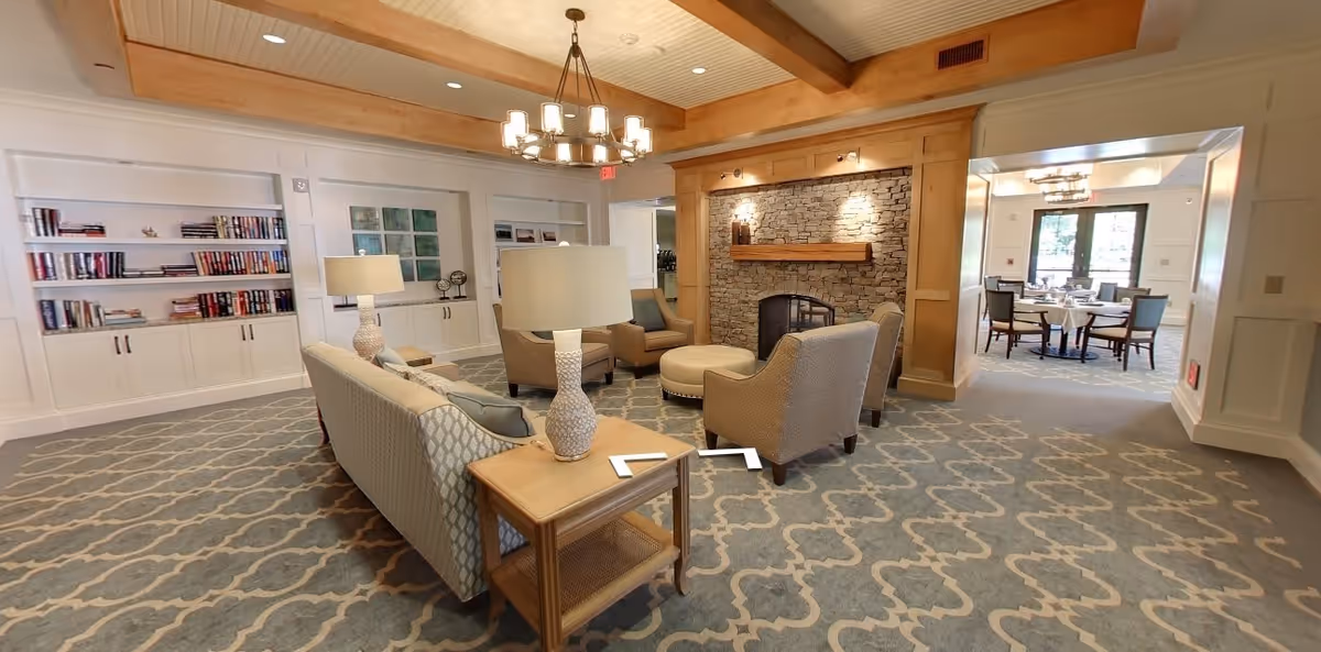 A spacious and well-lit living room area in a senior living facility featuring comfortable upholstered chairs and sofas arranged around a stone fireplace. The room has a patterned carpet, wooden ceiling beams, built-in bookshelves with books and decorative items, and a chandelier hanging from the ceiling. In the background, a dining area with tables and chairs is visible through a wide doorway.