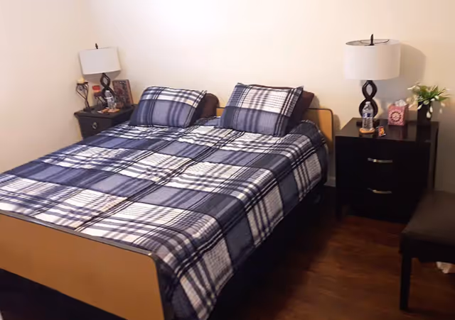 A bedroom with a double bed covered in a blue and white plaid bedspread and matching pillows. There are two black nightstands on either side of the bed, each with a lamp, and one nightstand has a water bottle, a box of tissues, and a small plant. The floor is wooden, and the walls are plain white.