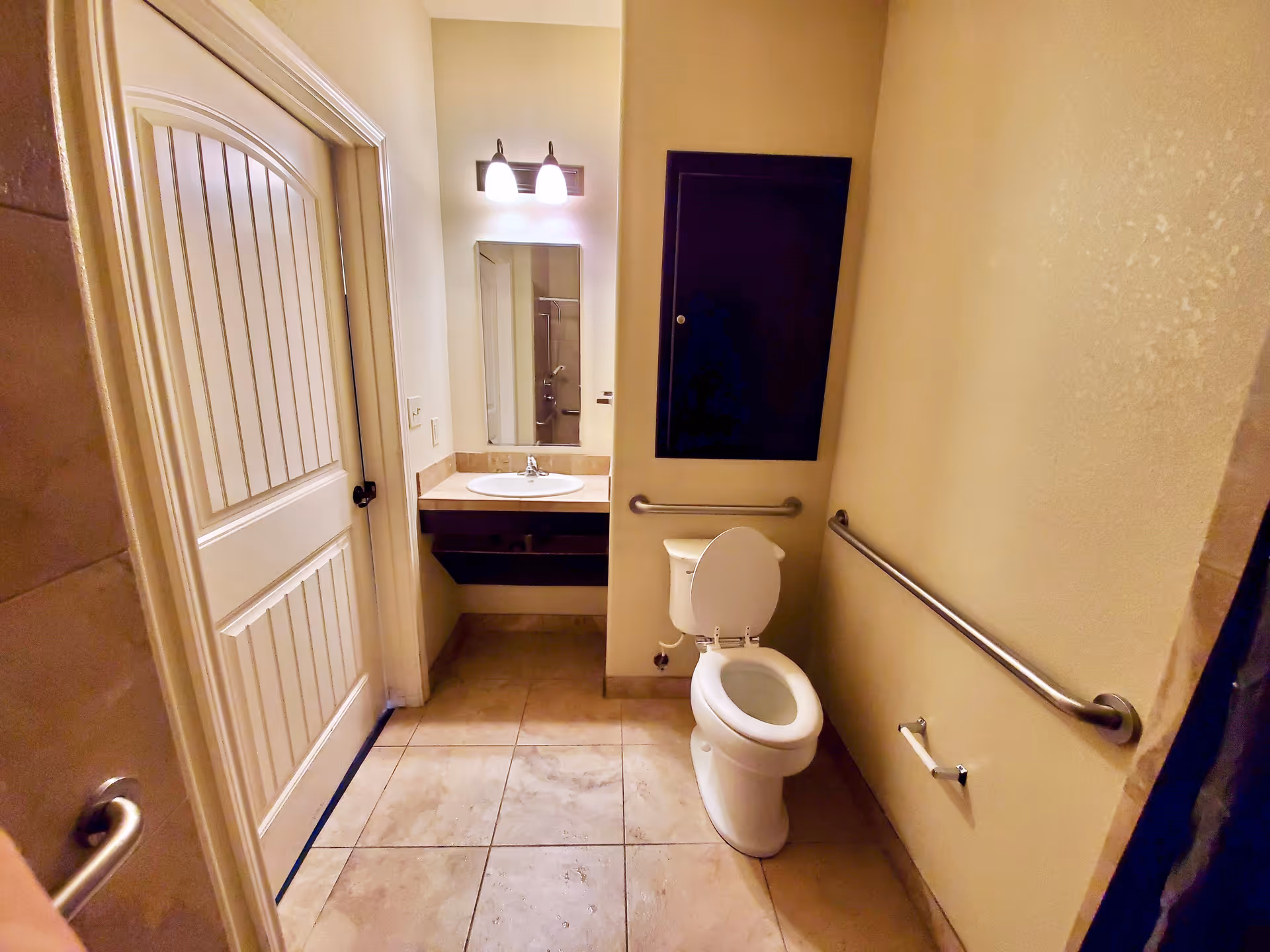 A small bathroom with beige walls and tiled floor. It features a white toilet with the lid open, a wall-mounted grab bar beside the toilet, a toilet paper holder, a sink with a countertop and a mirror above it, and a closed door on the left side. There is also a dark wall cabinet above the toilet and a two-light fixture above the mirror.