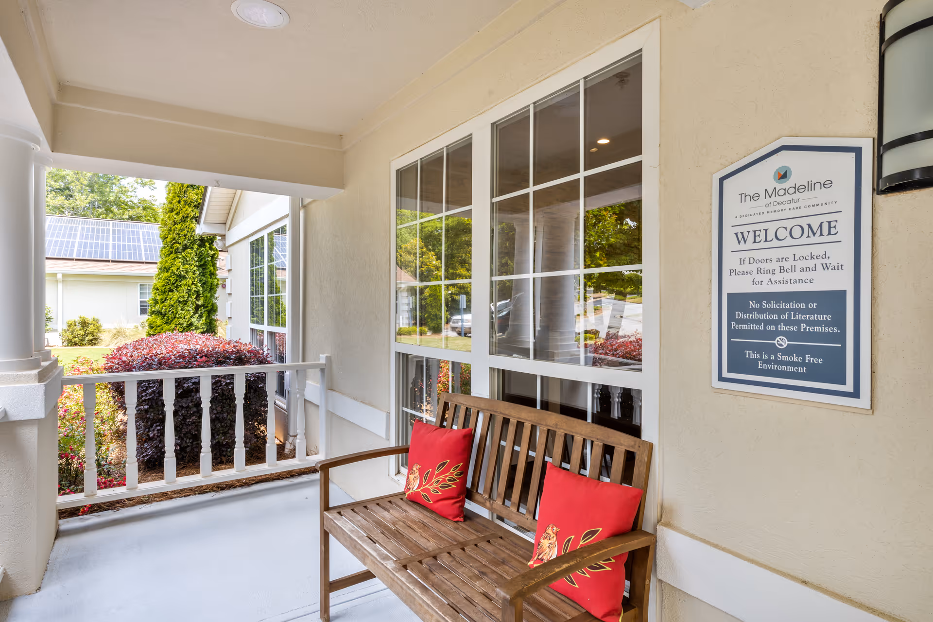Covered outdoor porch area with a wooden bench featuring two red decorative pillows. A large window is on the wall behind the bench, and a sign on the wall reads 'The Madeline of Decatur WELCOME If Doors are Locked, Please Ring Bell and Wait for Assistance No Solicitation or Distribution of Literature Permitted on these Premises This is a Smoke Free Environment'. Green shrubs and trees are visible outside.