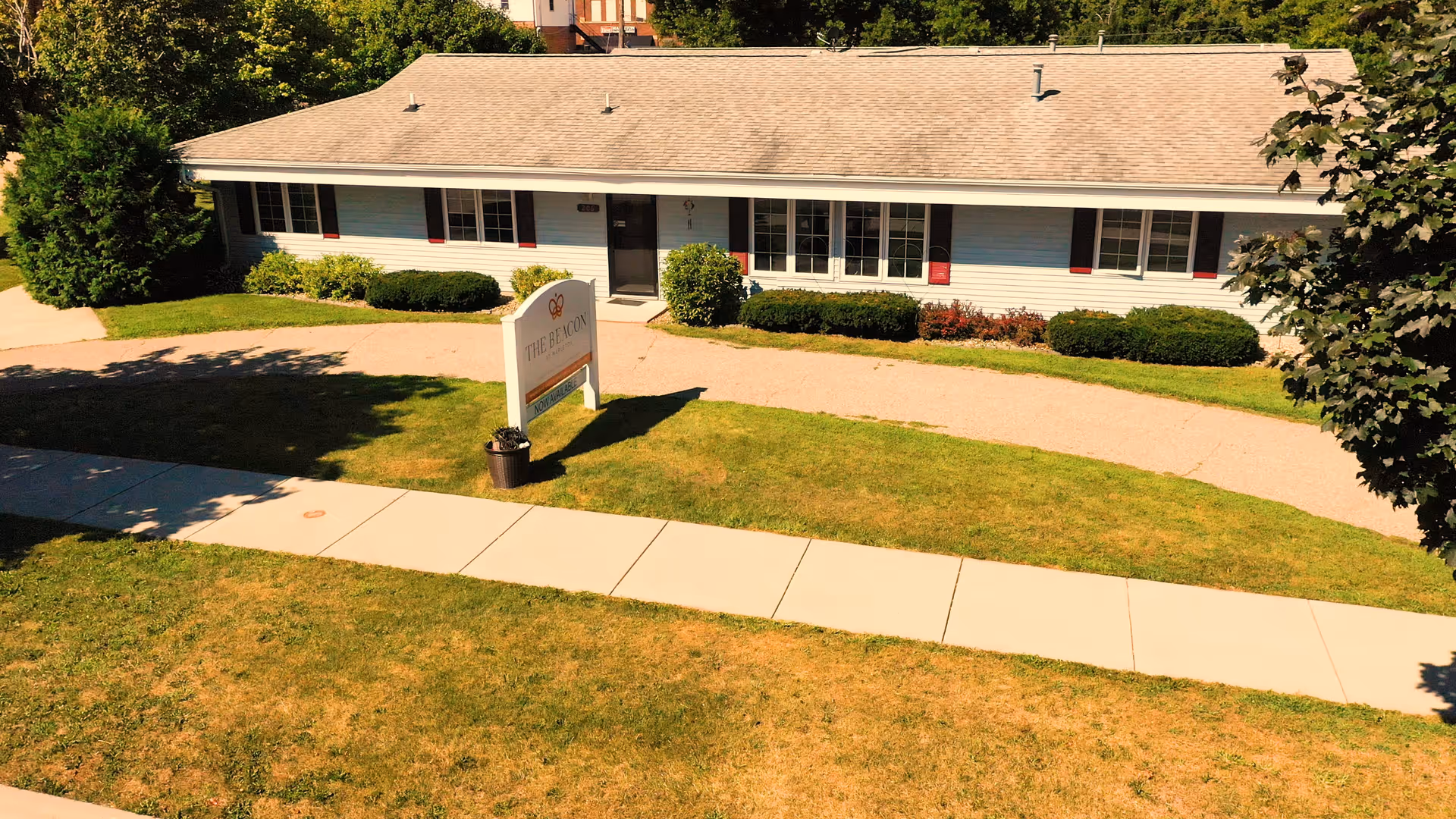 Single-story building with white siding and a gray shingled roof, surrounded by green bushes and trees. A sign in front reads 'The Beacon at Mapleton' with a smaller sign below stating 'Now Available.' A sidewalk runs in front of the building with a grassy lawn on either side.