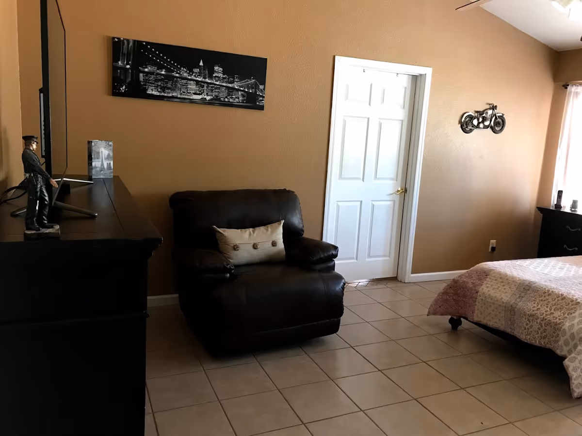 Bedroom with a dark leather armchair, dresser and TV, a bed, and a closed white door against tan walls.