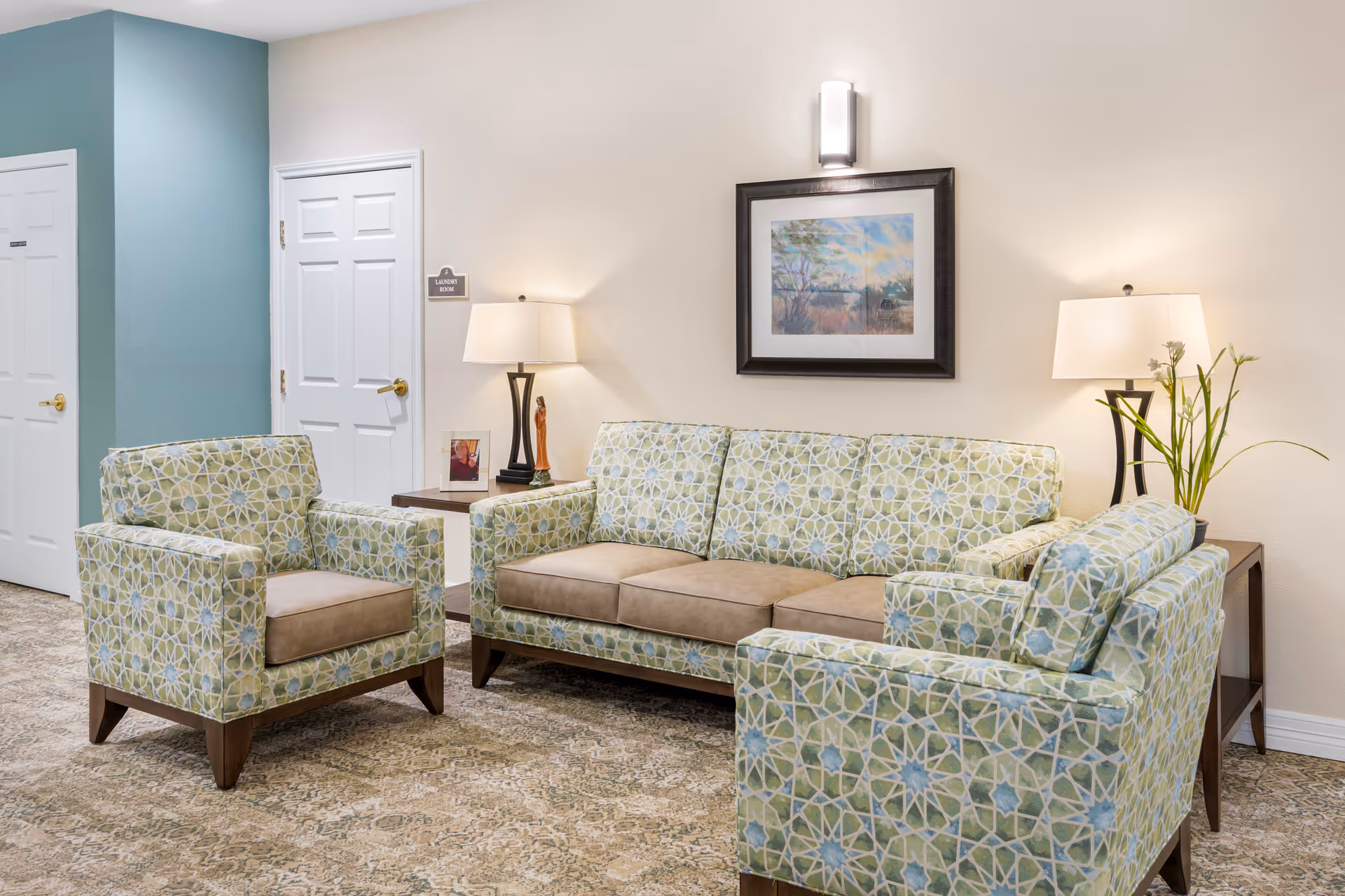 A bright lounge with patterned armchairs and a sofa, side tables with lamps, and framed artwork on the wall.