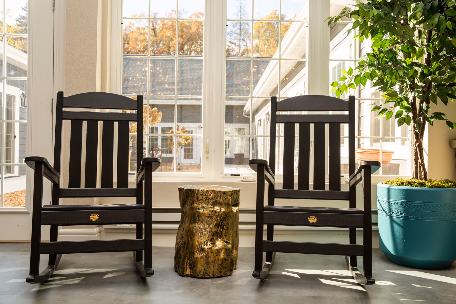 Two black wooden rocking chairs flanking a small tree-stump side table in front of large windows with a potted plant to the right.