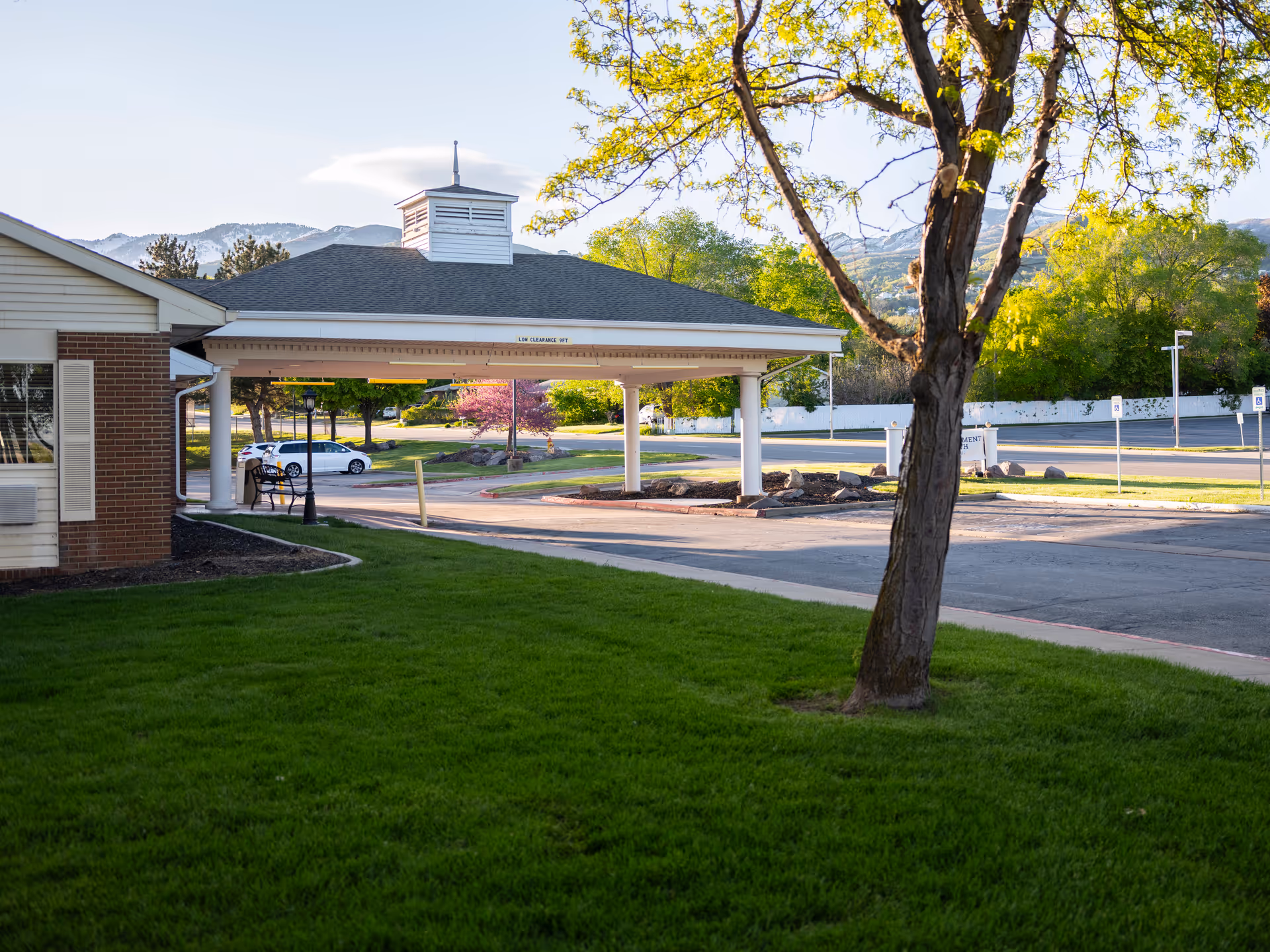 Outdoor view of the entrance area of Monument Healthcare Bountiful Skilled Nursing facility with a covered driveway, green lawn, a tree, and a parking lot in the background under a clear sky.