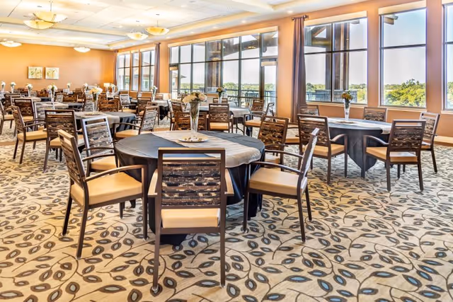 Spacious dining room with round tables and chairs, floral centerpieces, patterned carpet, and large windows overlooking greenery.