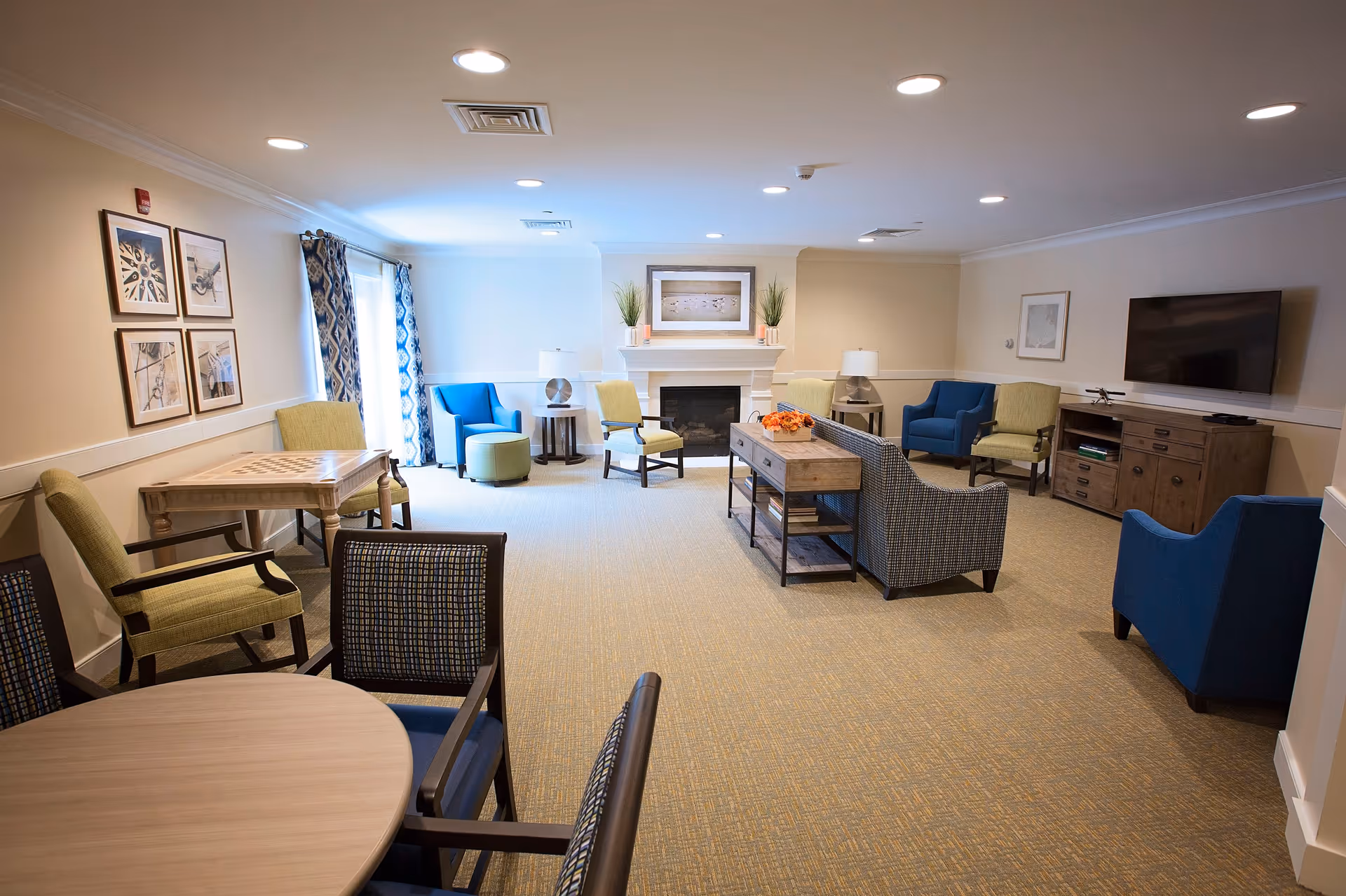 A spacious senior living common area with multiple seating options including armchairs, a sofa, and small tables. The room features a fireplace with decorative plants and a framed picture above it, a flat-screen TV mounted on the wall, and patterned curtains on the window. The carpet is beige, and the walls are painted light beige with white trim.