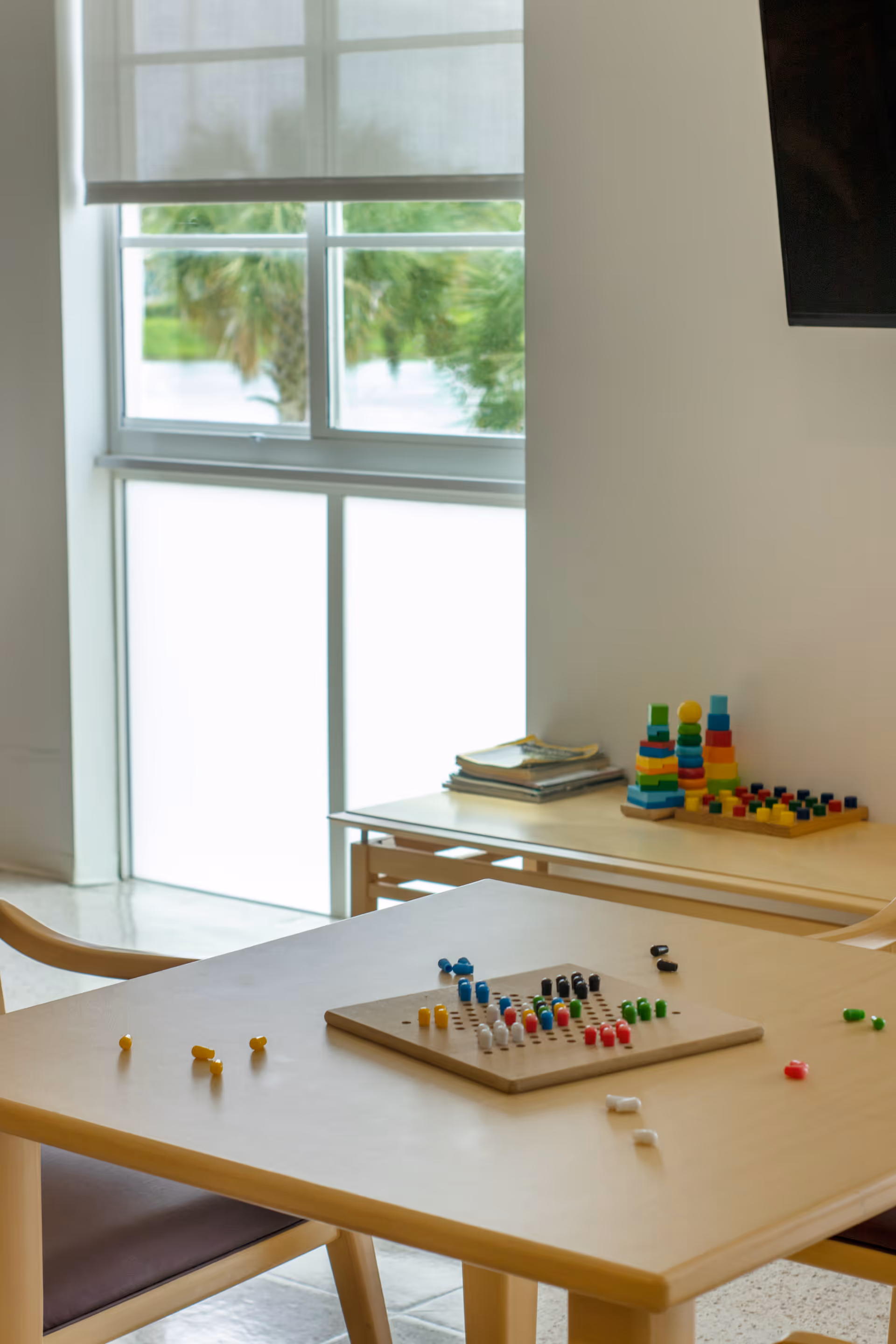 A light wooden table with a pegboard game featuring colorful pegs scattered on it, set in a bright room with large windows and a smaller table in the background holding stacked books and colorful stacking toys.