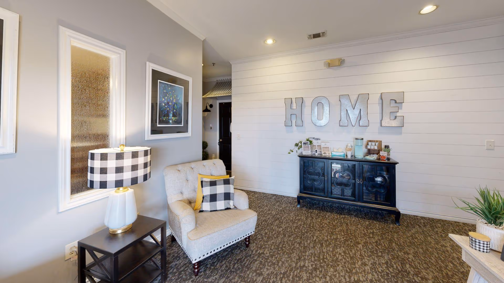 Comfortable seating area with an upholstered armchair, checkered lamp, black sideboard and large metal 'HOME' letters on a white shiplap wall.