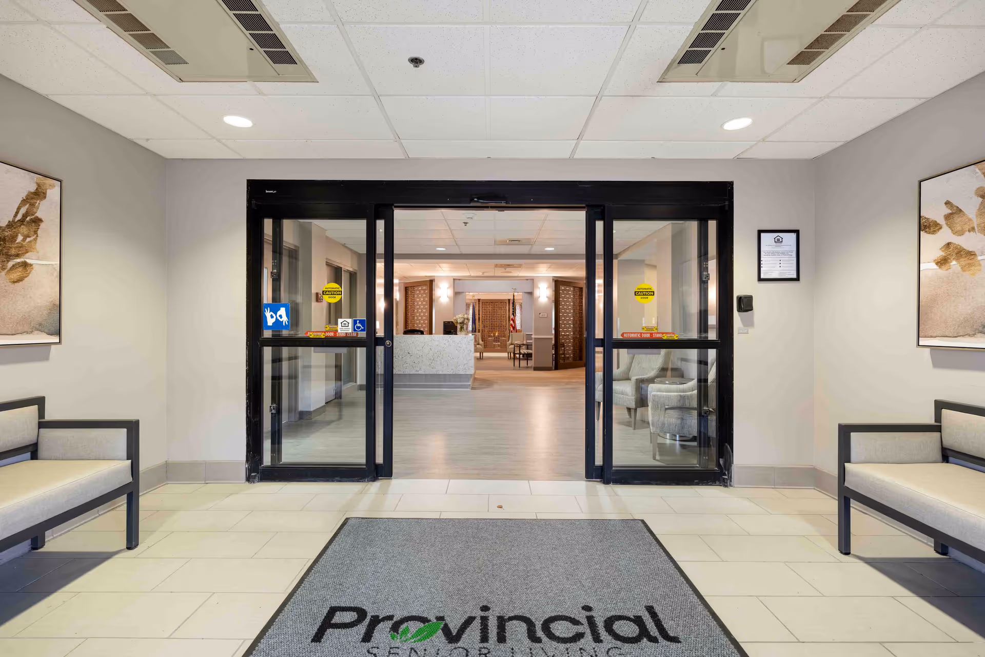 Entrance area of a senior living community with automatic sliding glass doors. Two benches with cushions are placed on either side of the entrance. There are framed abstract paintings on the walls above the benches. A gray floor mat with the word 'Provincial' is placed in front of the doors. Inside, a reception desk and seating area are visible.