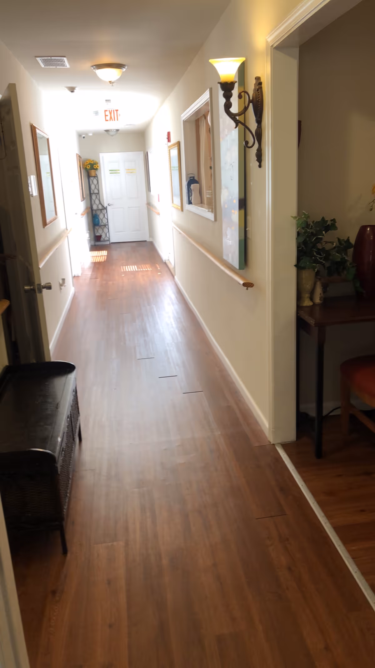 A well-lit hallway with wooden flooring and cream-colored walls. There are handrails along both sides of the hallway, framed pictures on the walls, and a decorative wall sconce light on the right. At the end of the hallway, there is a white door with an exit sign above it. On the right side near the foreground, there is a small table with plants and a red vase, and a chair partially visible.
